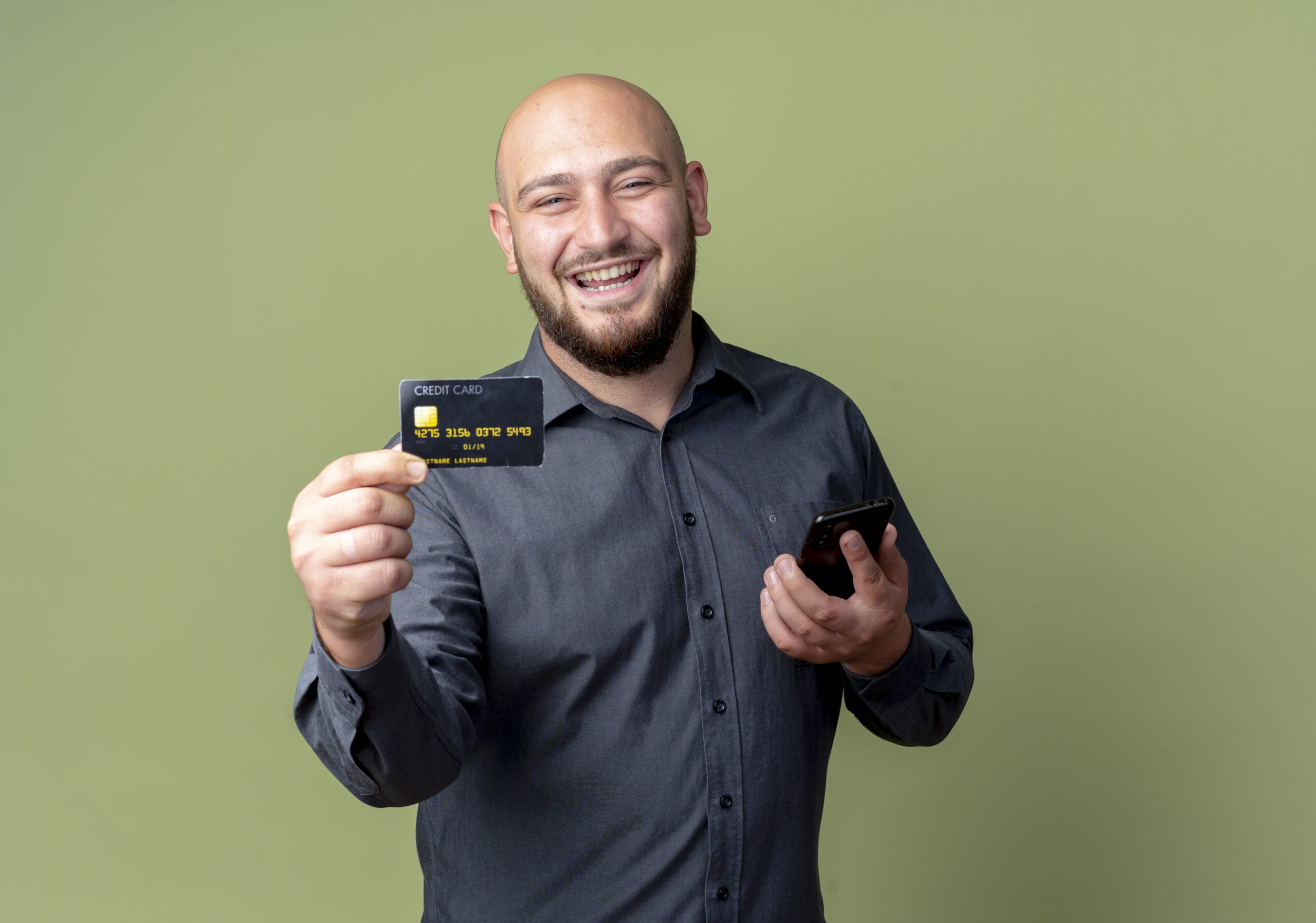 joyful young bald call center man holding cell phone and stretching out credit card at camera isolated on olive green background with copy space