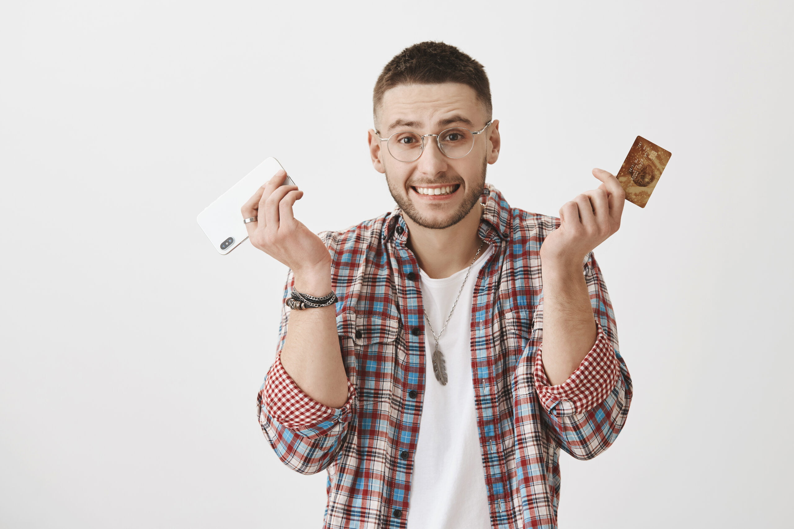 Siento haberme gastado todo tu dinero. Hombre torpe y apuesto encogiéndose de hombros y sosteniendo un smartphone y una tarjeta de crédito, sintiéndose incómodo mientras sonríe sobre un fondo gris. Uy, he comprado otro artículo en una tienda online.